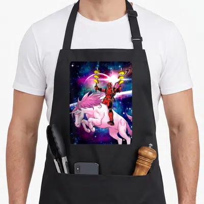 Space taco apron - Deadpool riding pink unicorn in space holding swords with three tacos each — shown on a black cotton canvas apron against a white background