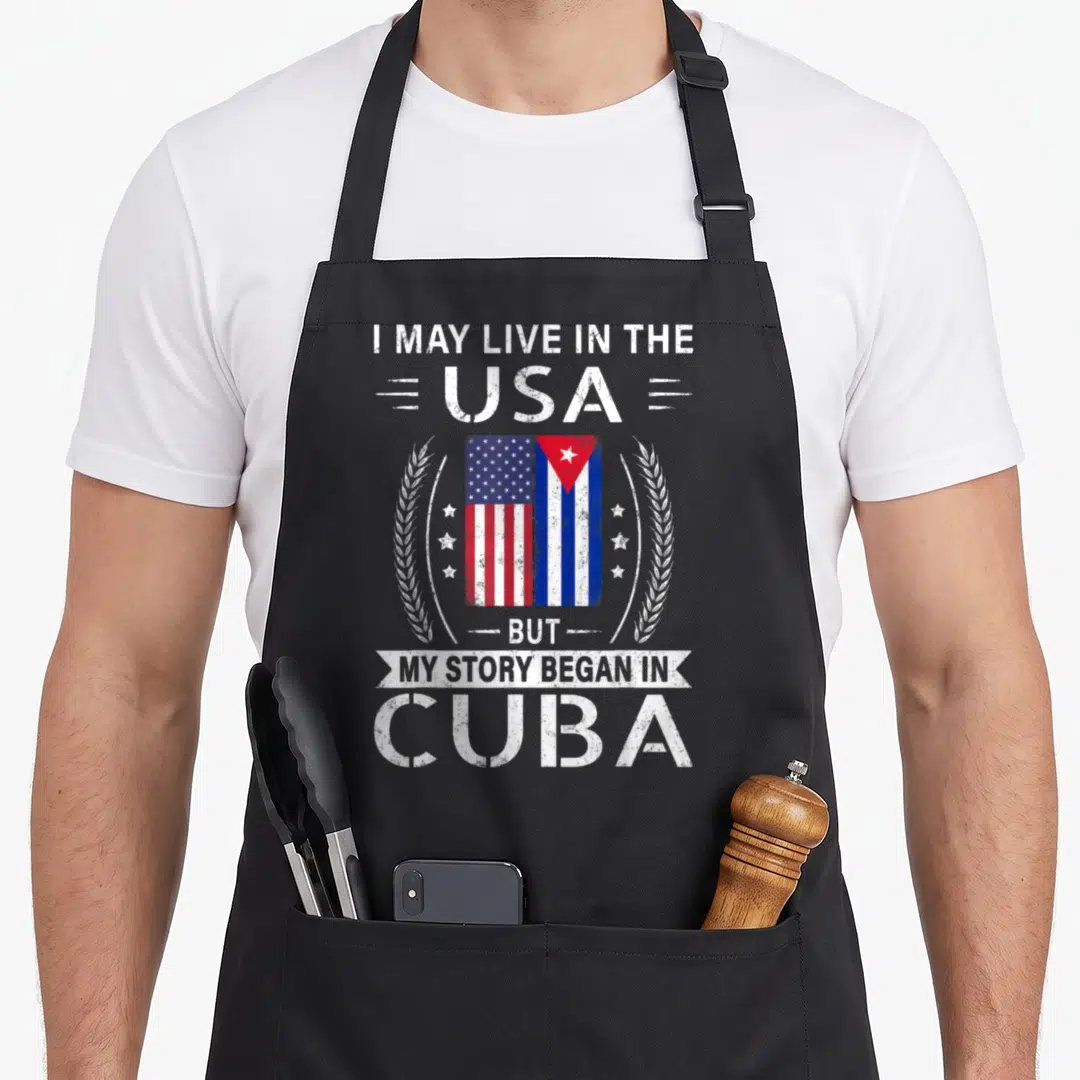 Cuban american flag apron - I MAY LIVE IN THE USA BUT MY STORY BEGAN IN CUBA split flag design with laurel wreath — shown on a black cotton canvas apron against a white background