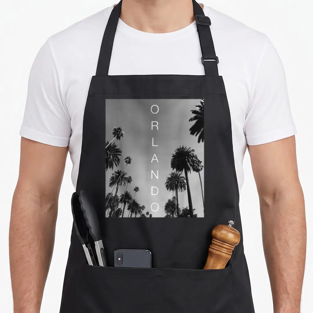 Orlando vacation florida apron - Vertical white ORLANDO text overlaid on black and white palm tree photo — shown on a black cotton canvas apron against a white background