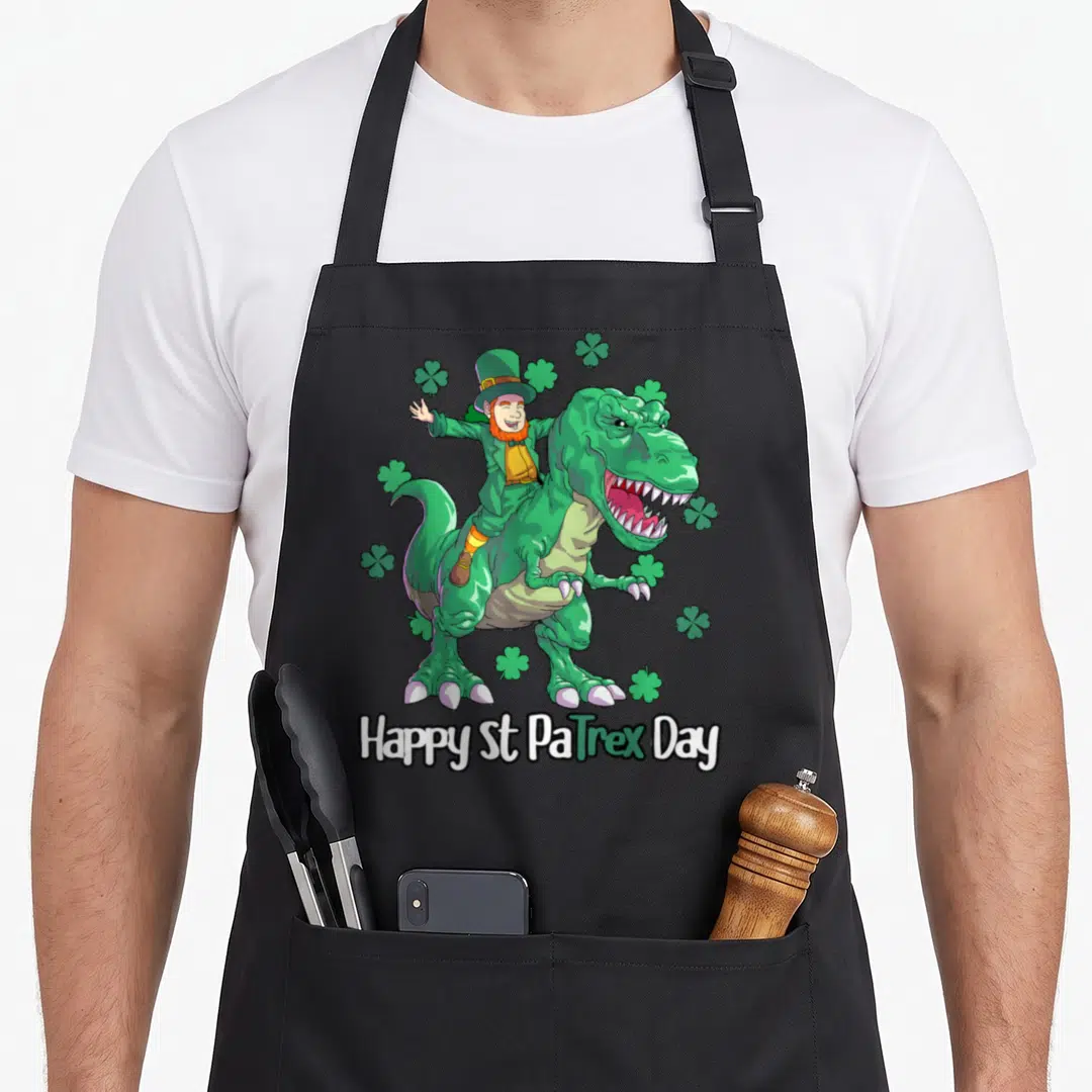 Dino st patrick day aprons - Leprechaun riding green T-Rex with Happy St PaTrex Day text and shamrocks — shown on a black cotton canvas apron against a white background