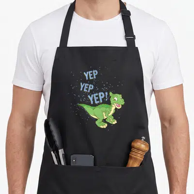 Land sprinkle apron - Green cartoon dinosaur with blue YEP YEP YEP text and scattered bubbles — shown on a black cotton canvas apron against a white background