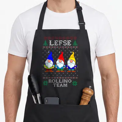 Lefse christmas baking team aprons - Three pixel art gnomes in blue, red, and yellow hats standing under LEPSE text — shown on a black cotton canvas apron against a white background
