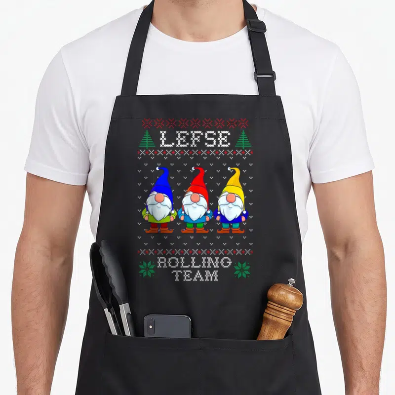 Lefse christmas baking team aprons - Three pixel art gnomes in blue, red, and yellow hats standing under LEPSE text — shown on a black cotton canvas apron against a white background