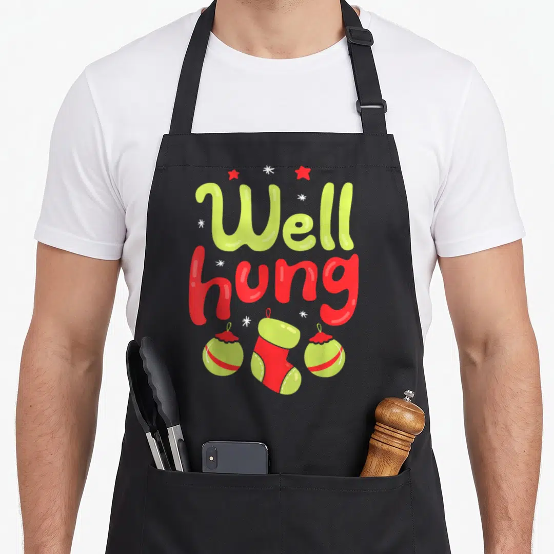 Hung christmas apron - Well hung text in lime and red with two ornaments and one stocking below — shown on a black cotton canvas apron against a white background