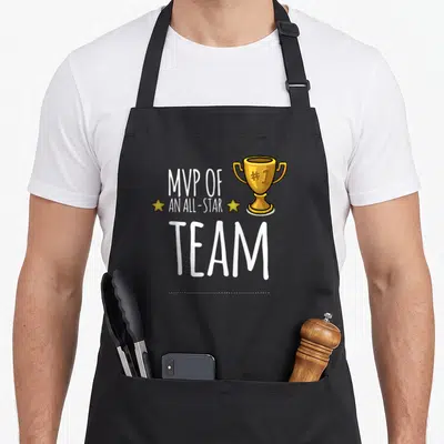 Mvp trophy apron - White MVP OF AN ALL-STAR TEAM text with gold trophy and two stars on black bib — shown on a black cotton canvas apron against a white background