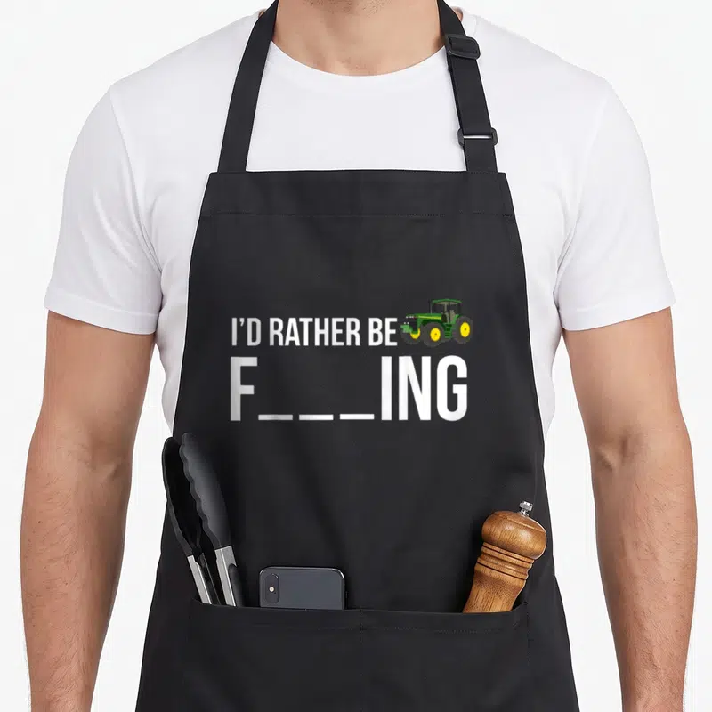 Rather farmer apron - I'd Rather Be tractor pun text with green and yellow vehicle illustration — shown on a black cotton canvas apron against a white background