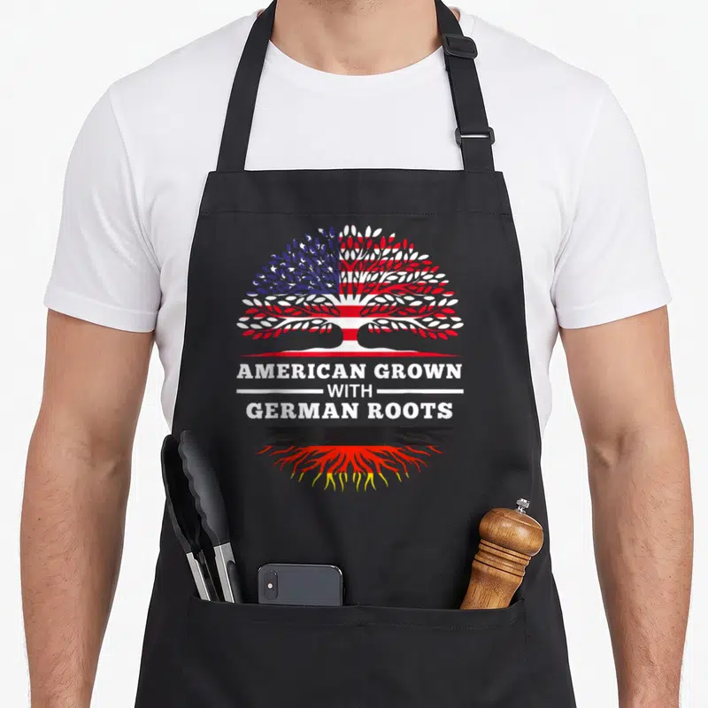 Root german apron - Tree with US flag leaves and German flag roots above AMERICAN GROWN WITH GERMAN ROOTS text — shown on a black cotton canvas apron against a white background