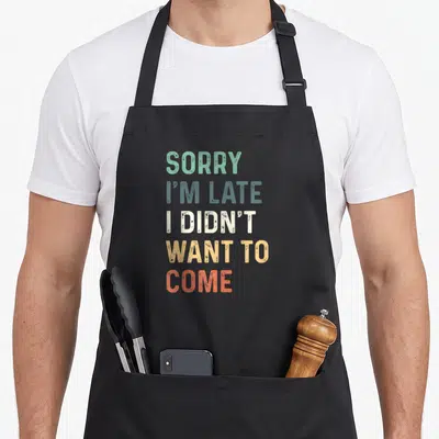 Didnt sorry im apron - Stacked SORRY I'M LATE text in green, blue-grey, white, yellow, and red-orange colors — shown on a black cotton canvas apron against a white background