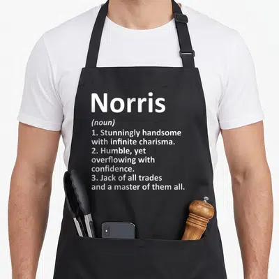 Norris father day aprons - White text defining Norris as handsome, humble, and a master of all trades on black fabric — shown on a black cotton canvas apron against a white background