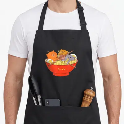 Cat kawaii aprons - Three cats in red ramen bowl with Japanese text and crossed chopsticks — shown on a black cotton canvas apron against a white background