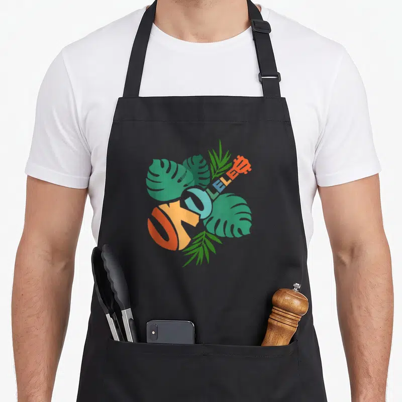 Hawaii ukulele apron - UKULELE text shaped like an instrument with green monstera leaves and orange blue accents — shown on a black cotton canvas apron against a white background