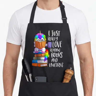 Bookworm reading book apron - Cartoon unicorn sitting on books holding a mug next to vertical text I just really love — shown on a black cotton canvas apron against a white background