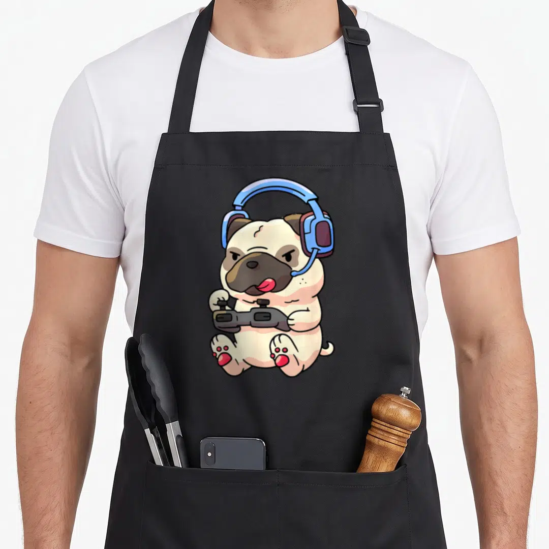 Pug video game apron - Cartoon beige pug sitting with blue headset and black game controller — shown on a black cotton canvas apron against a white background