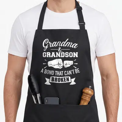Bond grandma apron - White distressed text Grandma Grandson with two fists bumping and broken ribbon banner — shown on a black cotton canvas apron against a white background