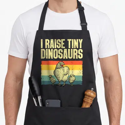 Chicken rooster hen apron - I RAISE TINY DINOSAURS text above hen and two chicks on striped background — shown on a black cotton canvas apron against a white background