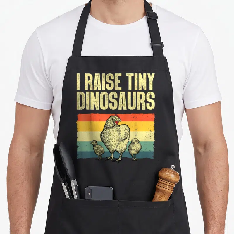 Chicken rooster hen apron - I RAISE TINY DINOSAURS text above hen and two chicks on striped background — shown on a black cotton canvas apron against a white background