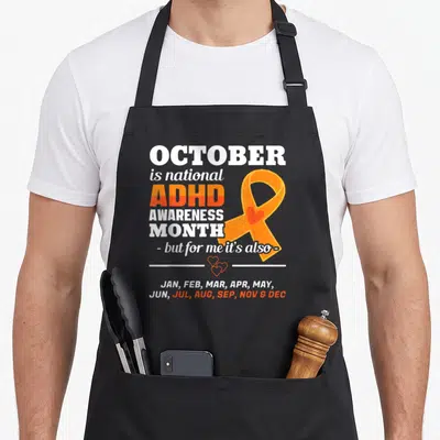 Adhd ribbon apron - Orange awareness ribbon with heart and stacked text listing all months for ADHD support — shown on a black cotton canvas apron against a white background