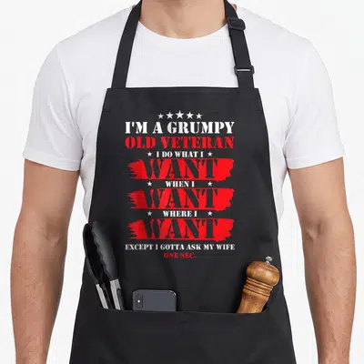 Grumpy veteran wife apron - Stacked white and red text reading I'm a grumpy old veteran with red brush strokes and stars — shown on a black cotton canvas apron against a white background