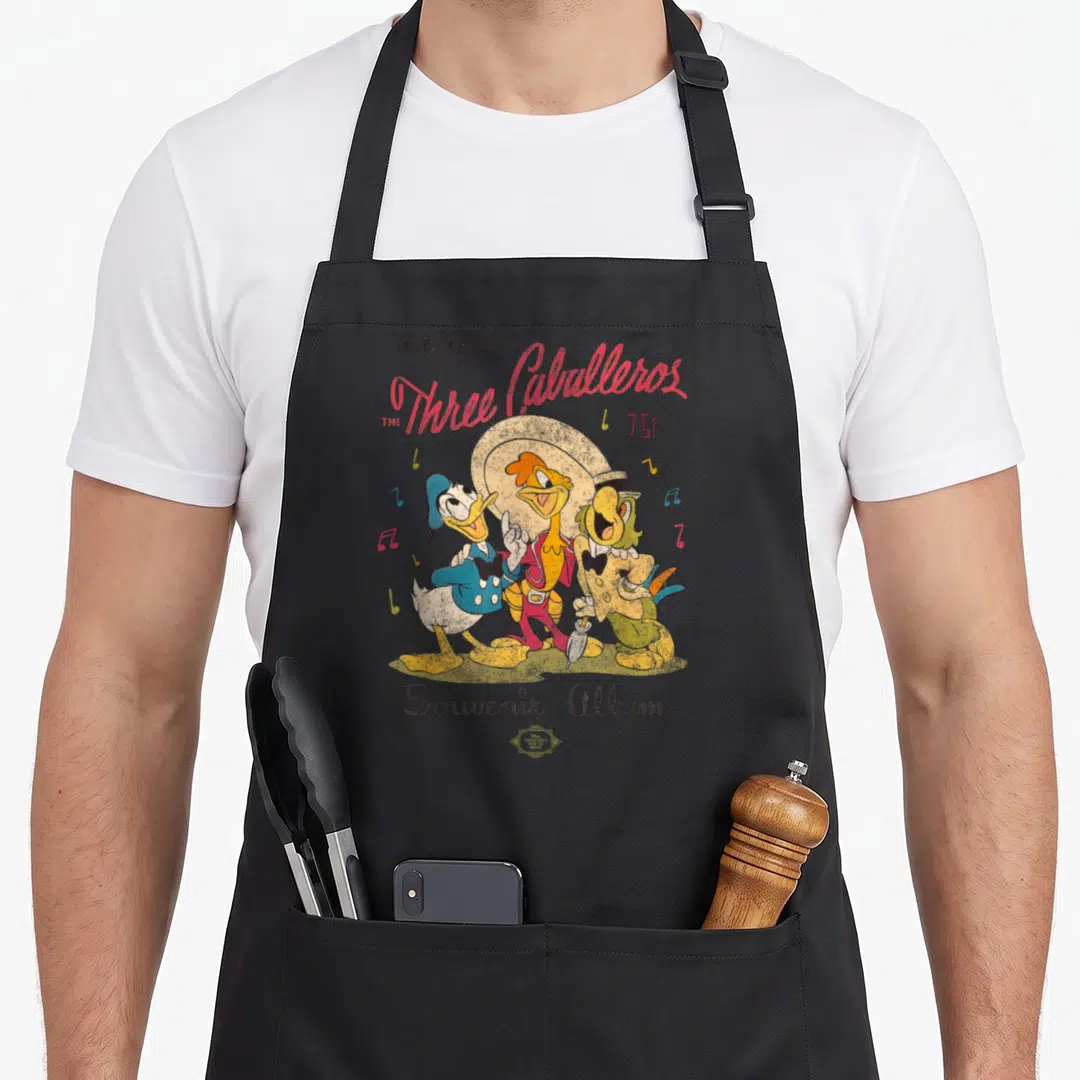 Caballero duck apron - Retro Three Caballeros cluster with stacked text and musical notes in red yellow blue green — shown on a black cotton canvas apron against a white background