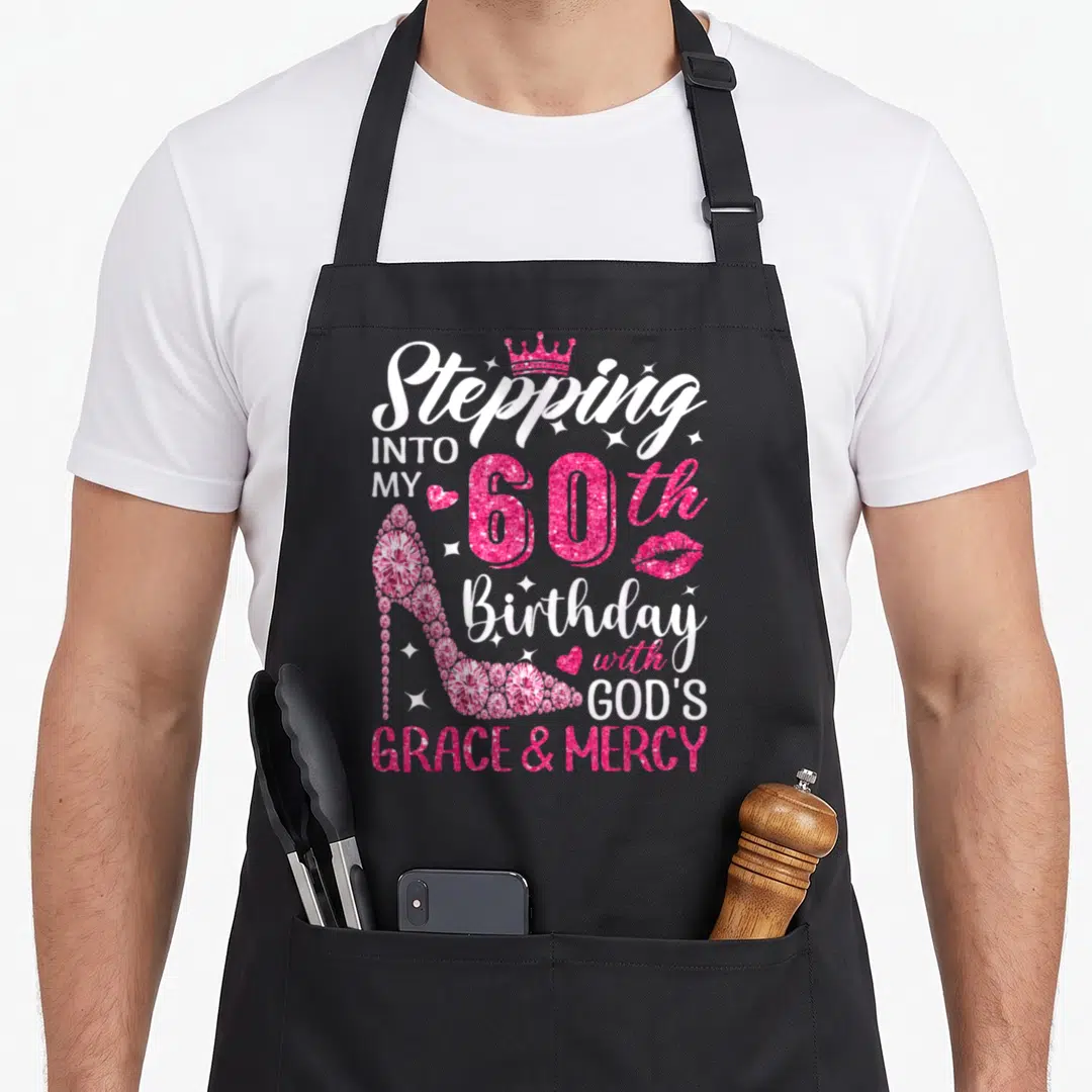 Grace 60th birthday apron - Pink glitter high heel shoe with crown and text Stepping Into My 60th Birthday with Gods — shown on a black cotton canvas apron against a white background