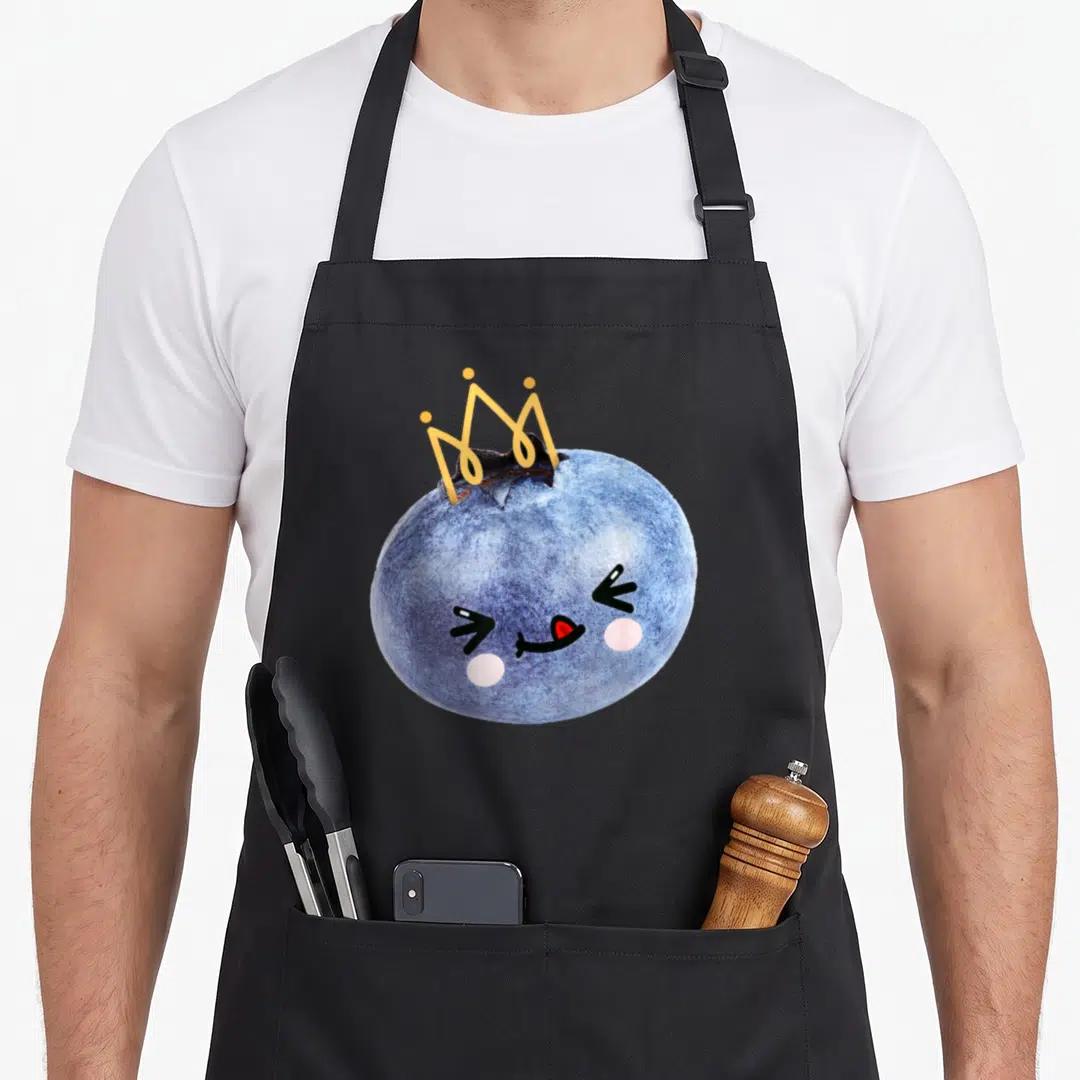 Blueberry anime apron - Cartoon blueberry with crown, pink cheeks, and yellow crown details — shown on a black cotton canvas apron against a white background