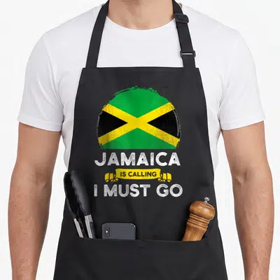 Jamaica heritage aprons - Distressed circular Jamaica flag above white text Jamaica Is Calling I Must Go with yellow — shown on a black cotton canvas apron against a white background
