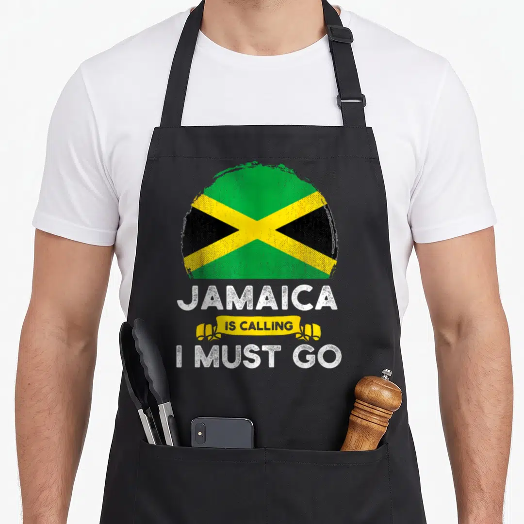 Jamaica heritage aprons - Distressed circular Jamaica flag above white text Jamaica Is Calling I Must Go with yellow — shown on a black cotton canvas apron against a white background