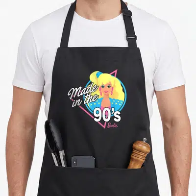 60th 90s apron - Cartoon woman portrait in blue circle and pink triangle with Made in the 90's Barbie text — shown on a black cotton canvas apron against a white background
