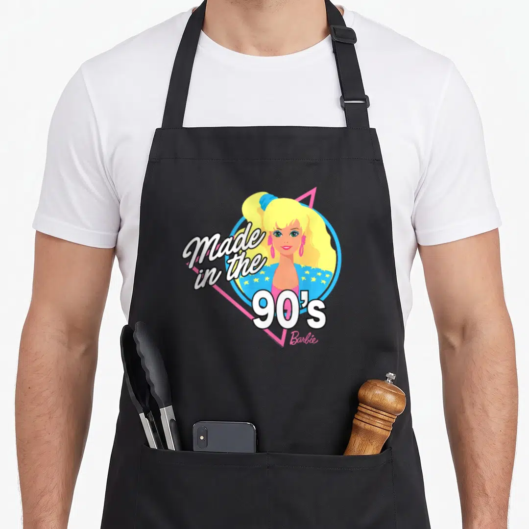 60th 90s apron - Cartoon woman portrait in blue circle and pink triangle with Made in the 90's Barbie text — shown on a black cotton canvas apron against a white background