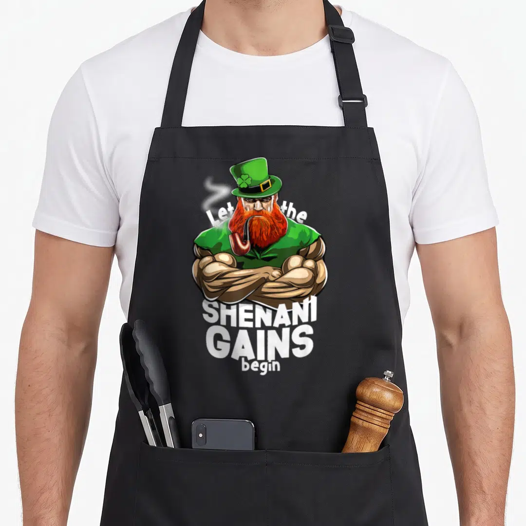 Jacked st patrick day aprons - Muscular leprechaun with crossed arms and text Let the SHENANI GAINS begin in green — shown on a black cotton canvas apron against a white background