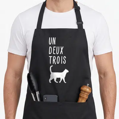 Deux french apron - White text UN DEUX TROIS above walking cat silhouette — shown on a black cotton canvas apron against a white background