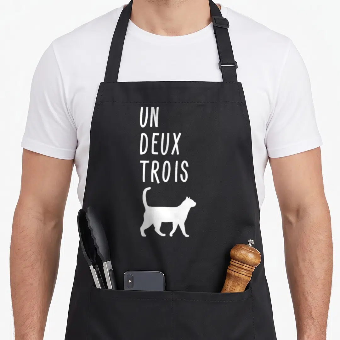 Deux french apron - White text UN DEUX TROIS above walking cat silhouette — shown on a black cotton canvas apron against a white background