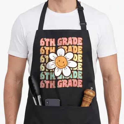 6th groovy grade apron - Seven multicolored rows of 6TH GRADE text behind a large smiling white daisy with small — shown on a black cotton canvas apron against a white background