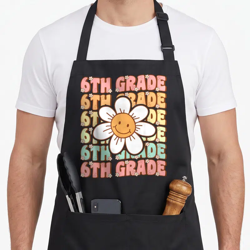 6th groovy grade apron - Seven multicolored rows of 6TH GRADE text behind a large smiling white daisy with small — shown on a black cotton canvas apron against a white background