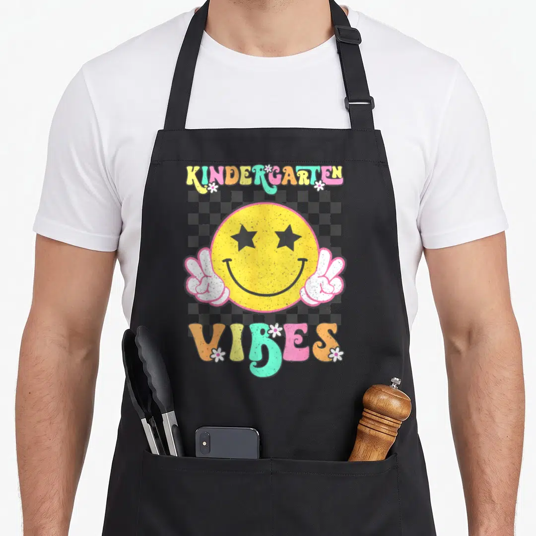 Kindergarten face apron - Yellow smiley face with star eyes and peace signs under multi-colored text on grey — shown on a black cotton canvas apron against a white background