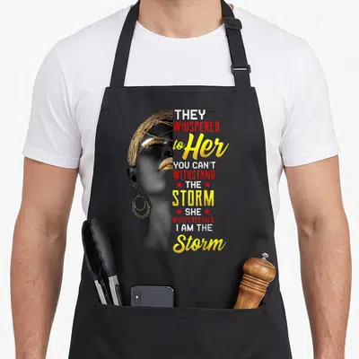 Strong queen african apron - Profile of woman with gold accessories and text THEY WHISPERED to Her I AM THE Storm — shown on a black cotton canvas apron against a white background