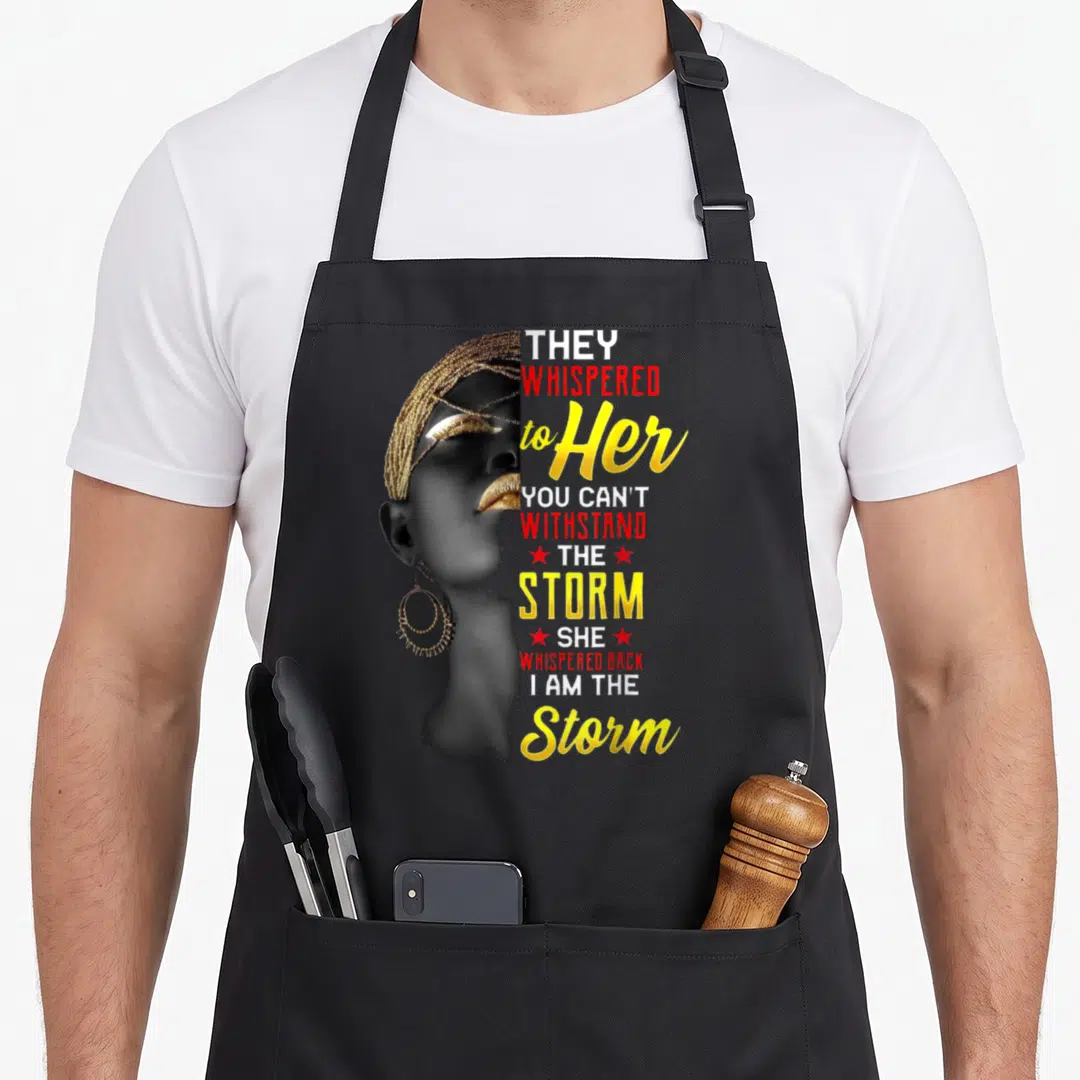 Strong queen african apron - Profile of woman with gold accessories and text THEY WHISPERED to Her I AM THE Storm — shown on a black cotton canvas apron against a white background