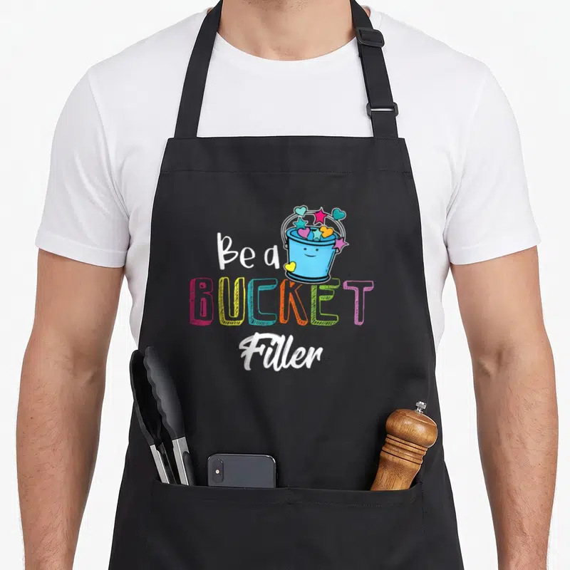 Filler teacher apron - Blue smiling bucket with hearts and stars next to Be a BUCKET Filler text — shown on a black cotton canvas apron against a white background