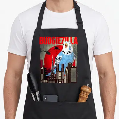 Budgiezilla bird apron - Giant blue budgie behind city skyline with red sun and BUDGIEZILLA text — shown on a black cotton canvas apron against a white background