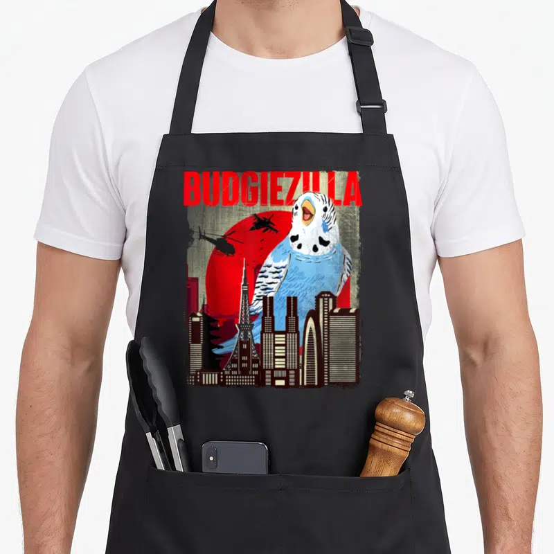 Budgiezilla bird apron - Giant blue budgie behind city skyline with red sun and BUDGIEZILLA text — shown on a black cotton canvas apron against a white background