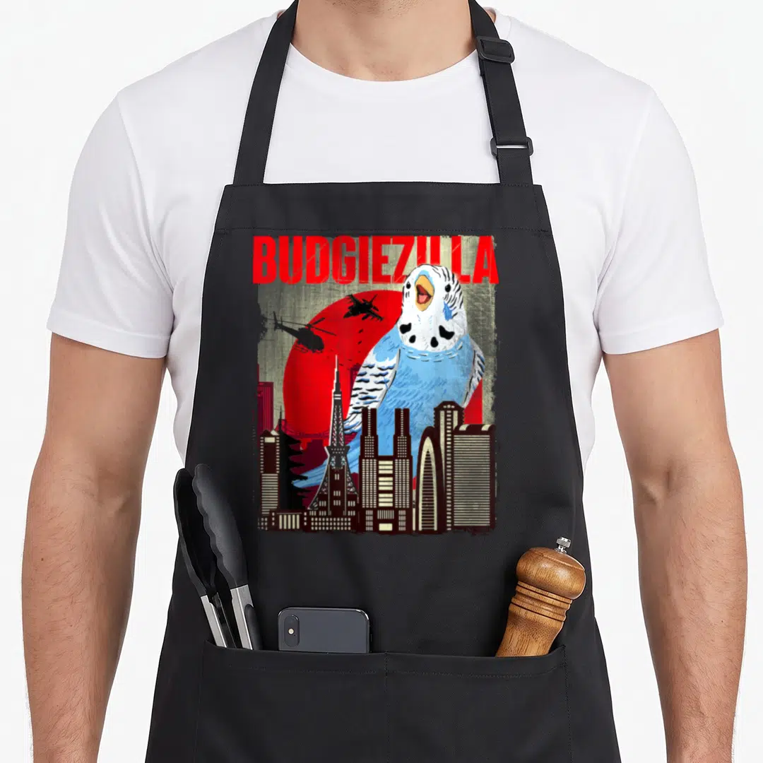 Budgiezilla bird apron - Giant blue budgie behind city skyline with red sun and BUDGIEZILLA text — shown on a black cotton canvas apron against a white background