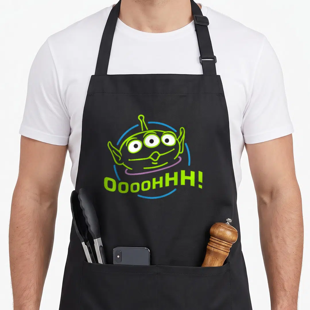 Funny alien apron - Lime green alien head with three eyes and OOOHHH text in blue and purple arcs — shown on a black cotton canvas apron against a white background