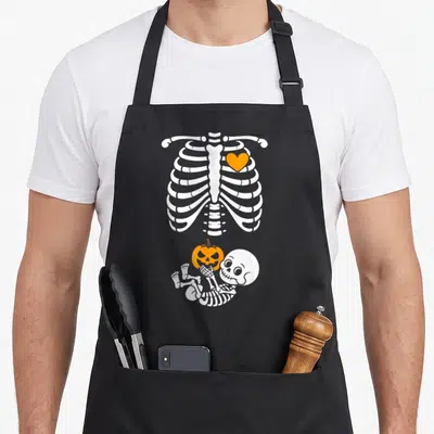 Baby halloween apron - White cartoon ribcage with orange heart above a lying baby skeleton holding an orange pumpkin — shown on a black cotton canvas apron against a white background