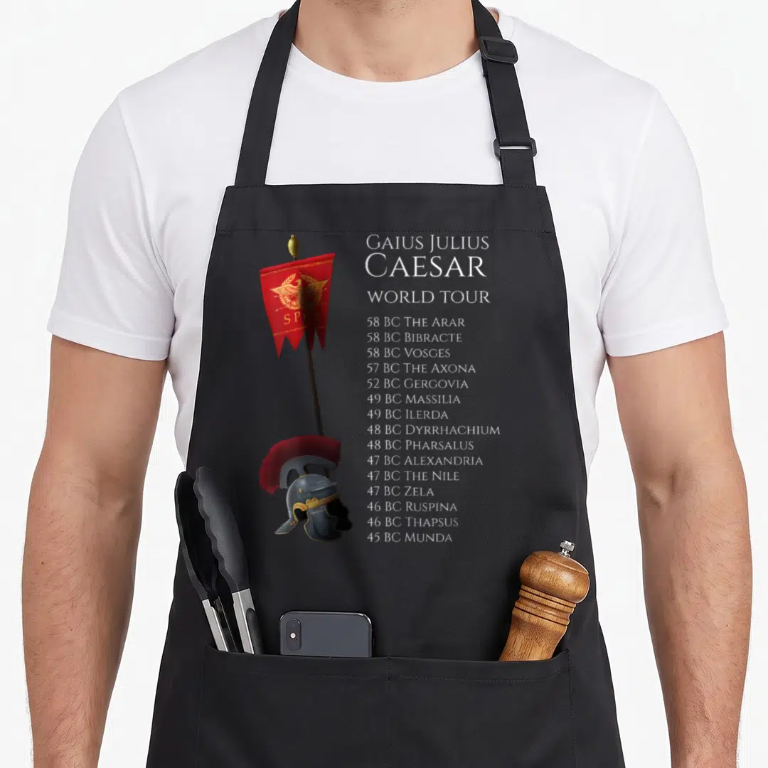 Ancient bloody caesar apron - Roman standard helmet and Caesar world tour battle list in red gold black white — shown on a black cotton canvas apron against a white background