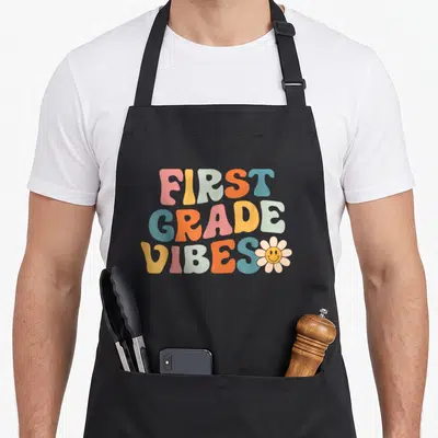 1st grade vibes apron - Stacked retro text reading FIRST GRADE VIBES in pink yellow orange and blue with a smiley flower — shown on a black cotton canvas apron against a white background