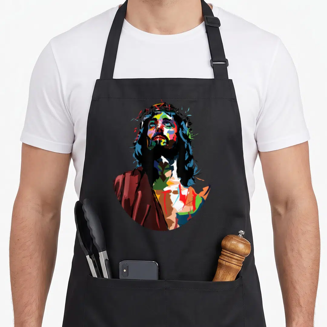 God king of the grill apron - Multicolored cartoon portrait of Jesus wearing a crown of thorns in blue red yellow green — shown on a black cotton canvas apron against a white background