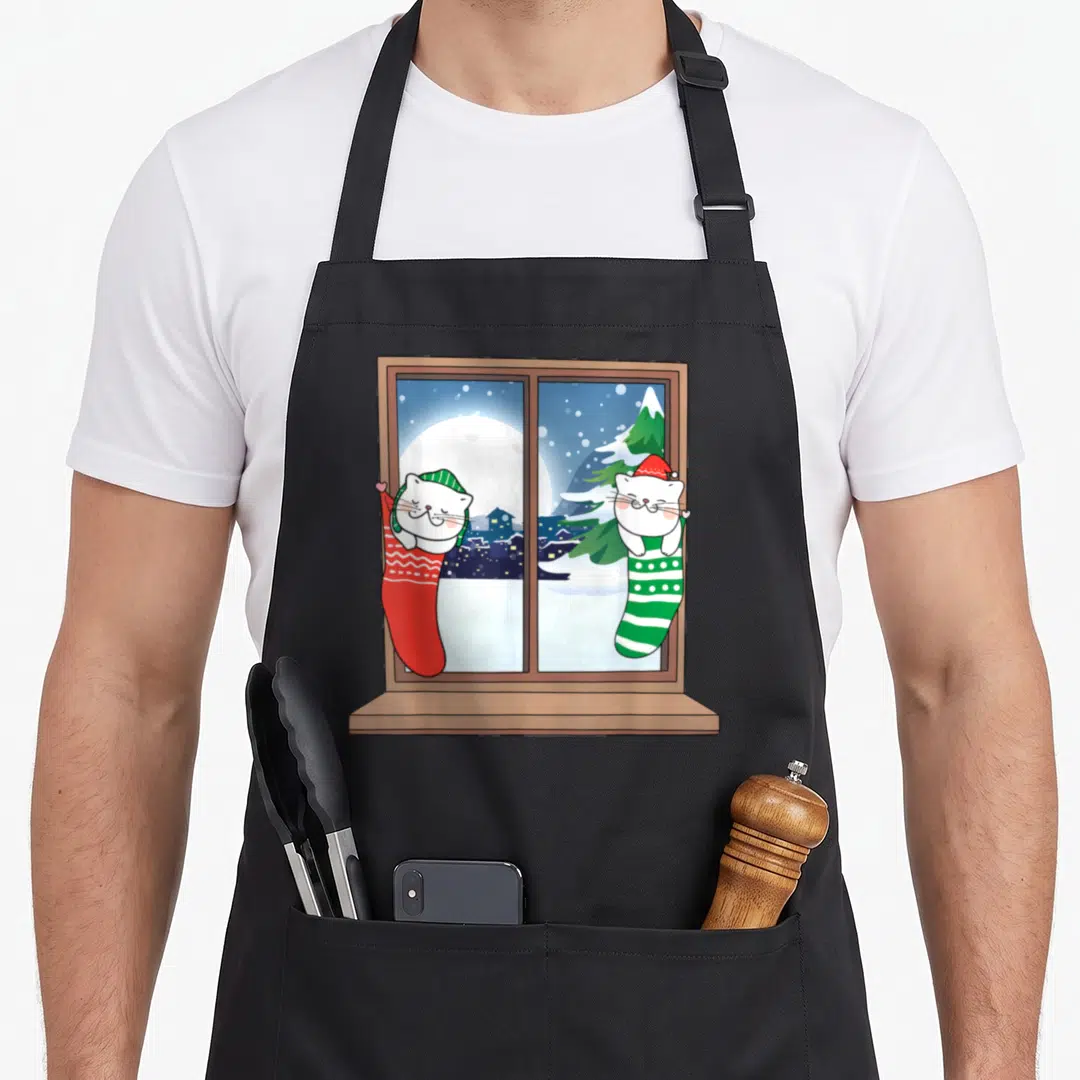Twin womens christmas aprons - Two white cats in red and green stockings inside a window frame with a snowy night scene — shown on a black cotton canvas apron against a white background