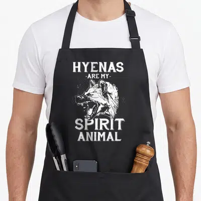 Africa animal apron - White hyena head illustration with halftone dots and distressed text reading HYENAS ARE MY SPIRIT — shown on a black cotton canvas apron against a white background