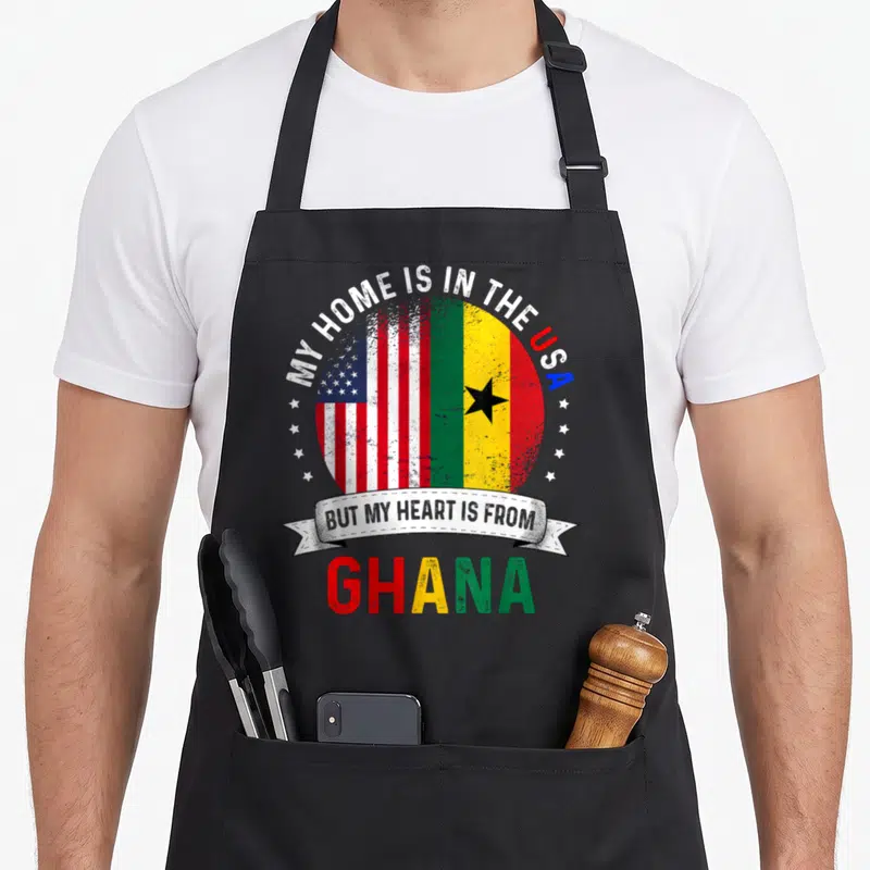Ghana patriots apron - Split USA and Ghana flags with text My Home Is In The USA But My Heart Is From Ghana — shown on a black cotton canvas apron against a white background