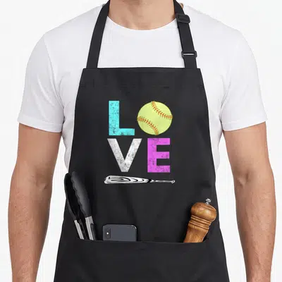 Softball birthday apron - LOVE spelled with blue L, yellow softball, white V, pink E, and horizontal bat — shown on a black cotton canvas apron against a white background